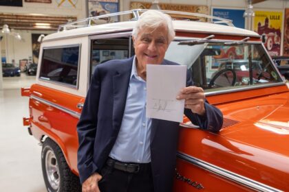Jay Leno holding signed paper beside orange classic Ford Bronco